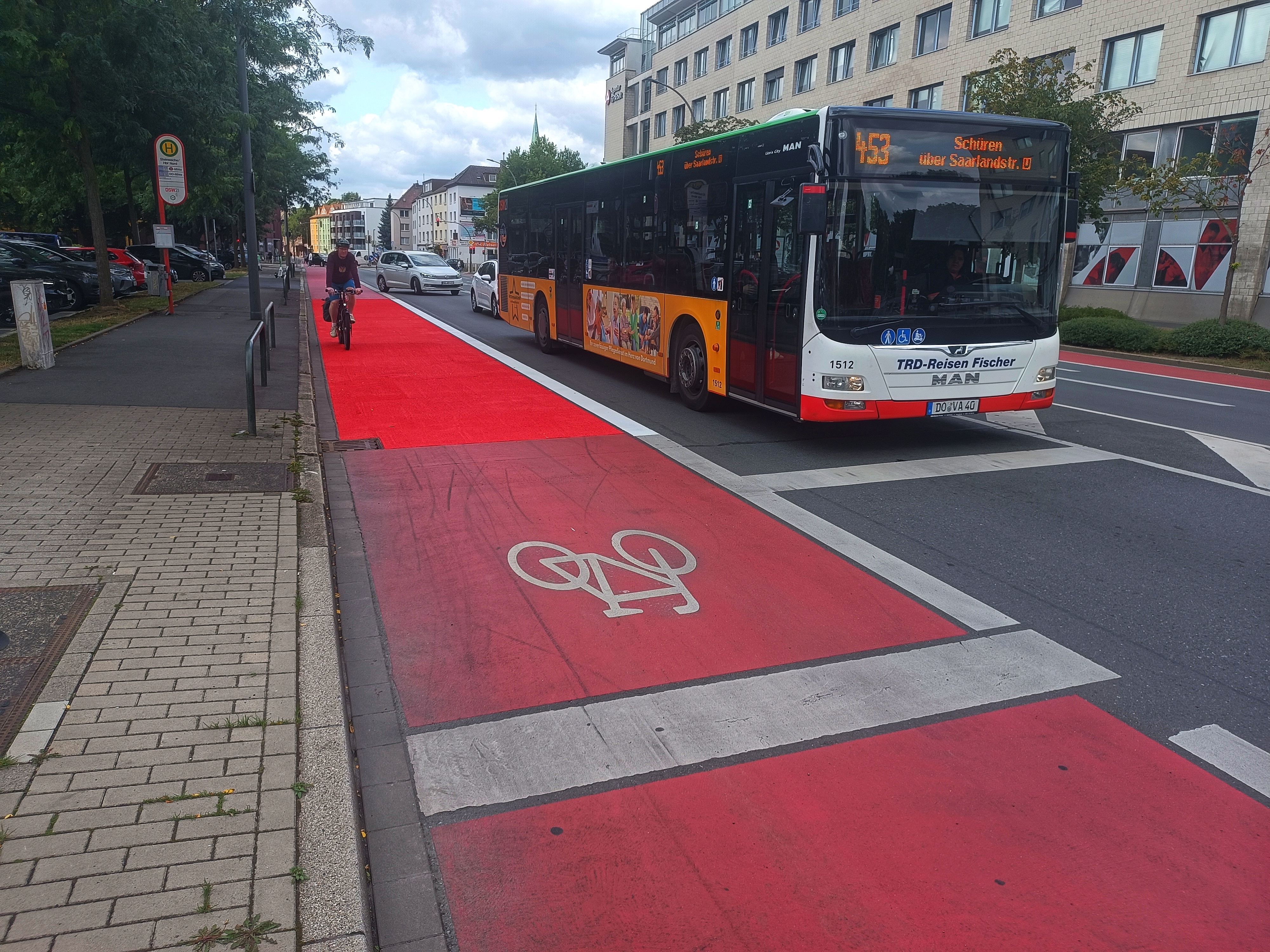 Ein roter Radfahrstreifen. Ein Teilstück wurde neu markiert. Die Farbe leuchtet heller als auf den älteren Abschnitten des Weges. Recht im Bild wartet ein Bus vor der Ampel. Links radelt ein Radfahrer über den neuen Radfahrstreifen. 