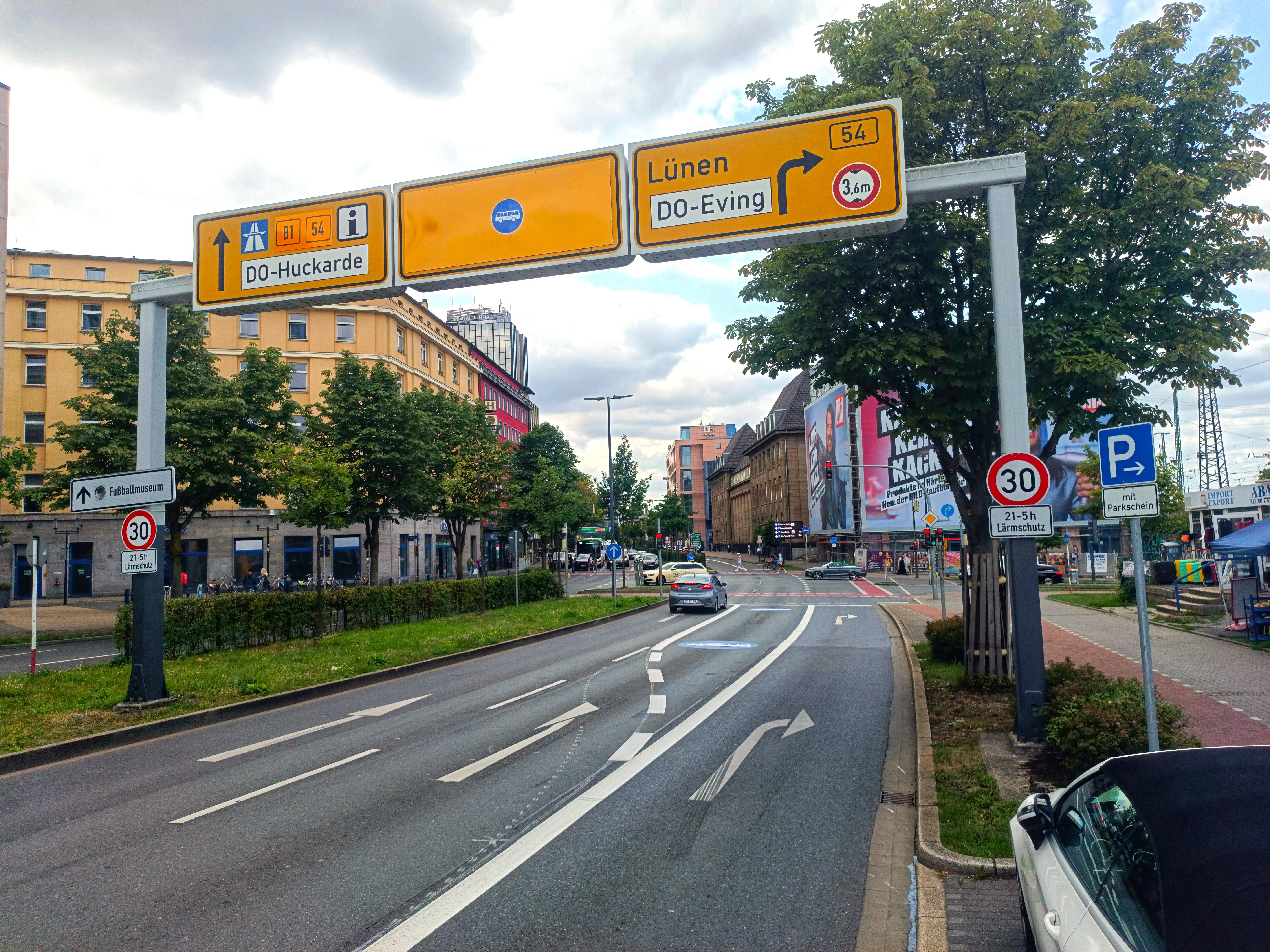 Straßenansicht in einer städtischen Kulisse. Drei Fahrspuren liegen nebeneinander. In der mittleren Fahrspur beginnt eine Busspur. Im Hintergrund es eine rote Ampel an der nächsten Kreuzung zu sehen. Im Vordergrund ist eine Schilderbrücke mit drei gelben Schildern zu sehen. Weitere Wegweiser, Verkehrs- und Parkschilder stehen am linken und rechten Bildrand. 