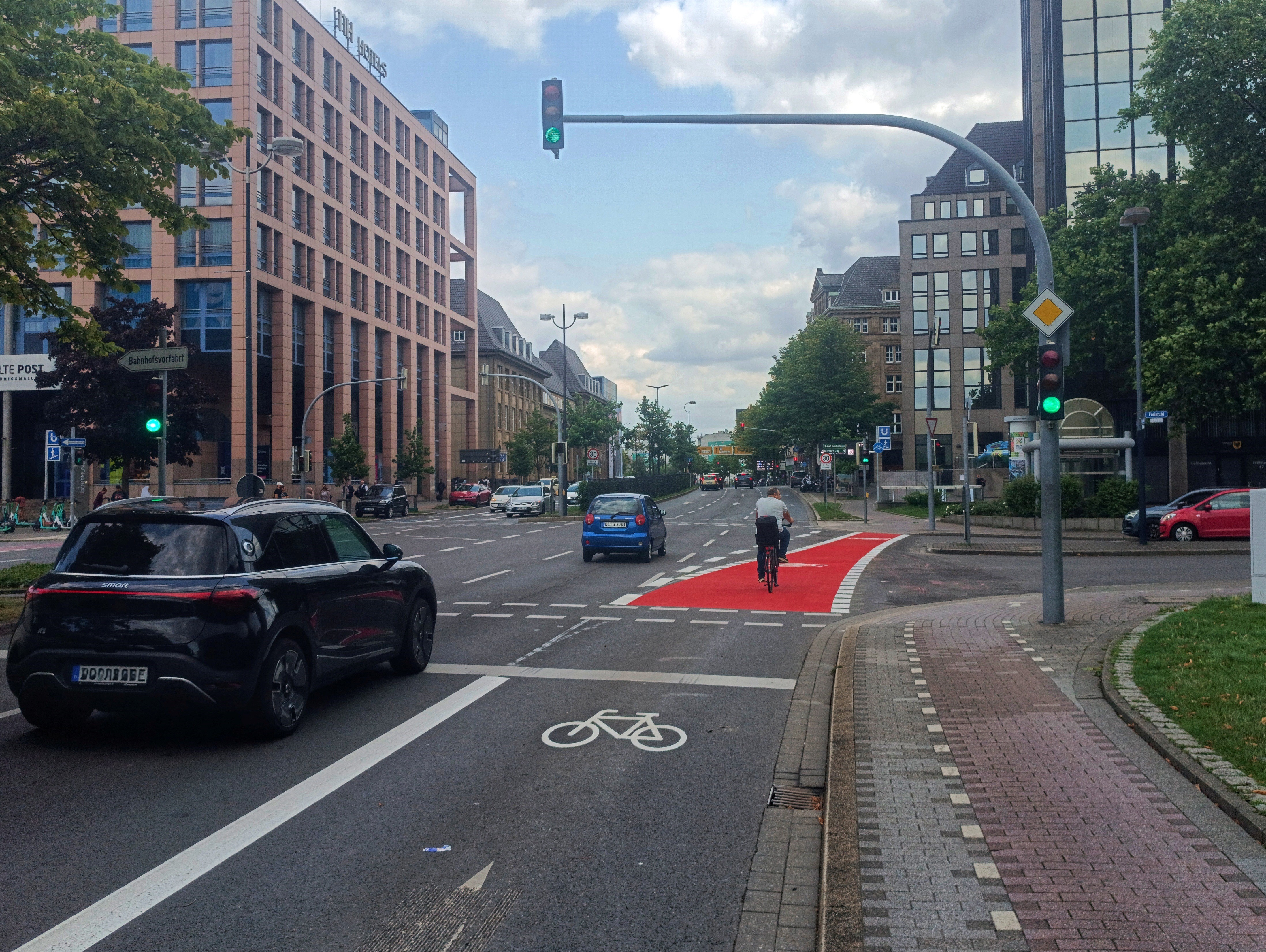 Städtische Kreuzung mit mehreren Fahrspuren und Radwegmarkierungen. Im Vordergrund ist ein weißes Fahrradsymbol auf der Fahrbahn zu sehen. Rechts daneben verläuft ein abgesetzter Bürgersteig. Links fährt ein schwarzes Auto geradeaus, im Hintergrund sind weitere Autos und ein mehrstöckiges Gebäude aus rotem Stein sowie moderne Glasgebäude zu erkennen. Die Ampeln zeigen für Autos und Radfahrer Grün. Links am Straßenrand stehen E-Scooter und Verkehrsschilder, im Hintergrund Bäume und Straßenschilder.