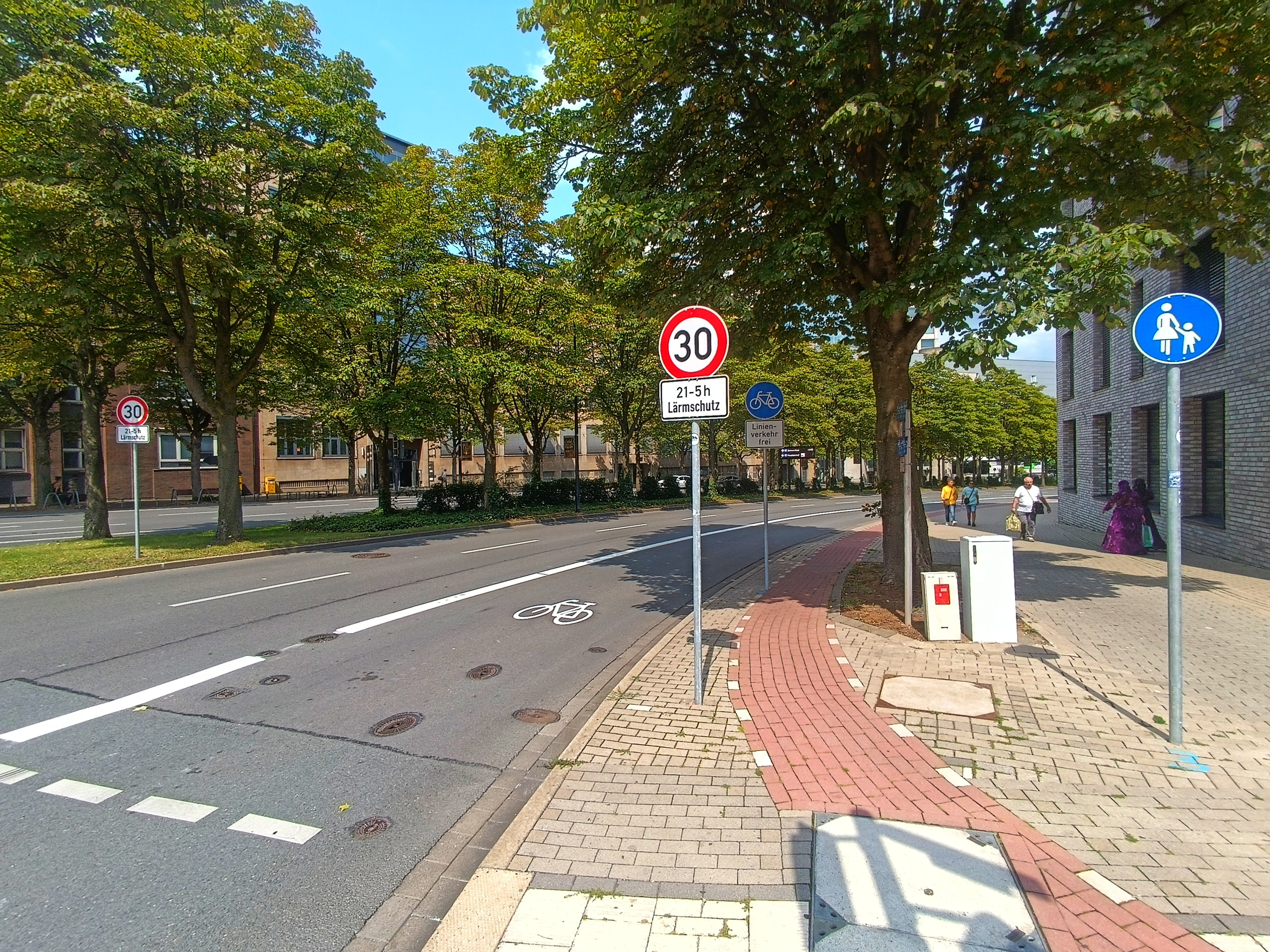 Straßenansicht in der Stadt. Links sind zwei Fahrspuren für Autos zu sehen. Daneben ist ein Radfahrstreifen mit Fahrradpiktogramm zu sehen. Der Bürgersteig ist als reiner Gehweg ausgeschildert. In der Mitte des Bildes ist ein Tempo 30 Schild zu sehen. Der Straßenverlauf wird von Bäumen begleitet. Links im Bild gehen Fußgägner*innen. Es ist sommerlich und der Himmel wolkenlos blau.