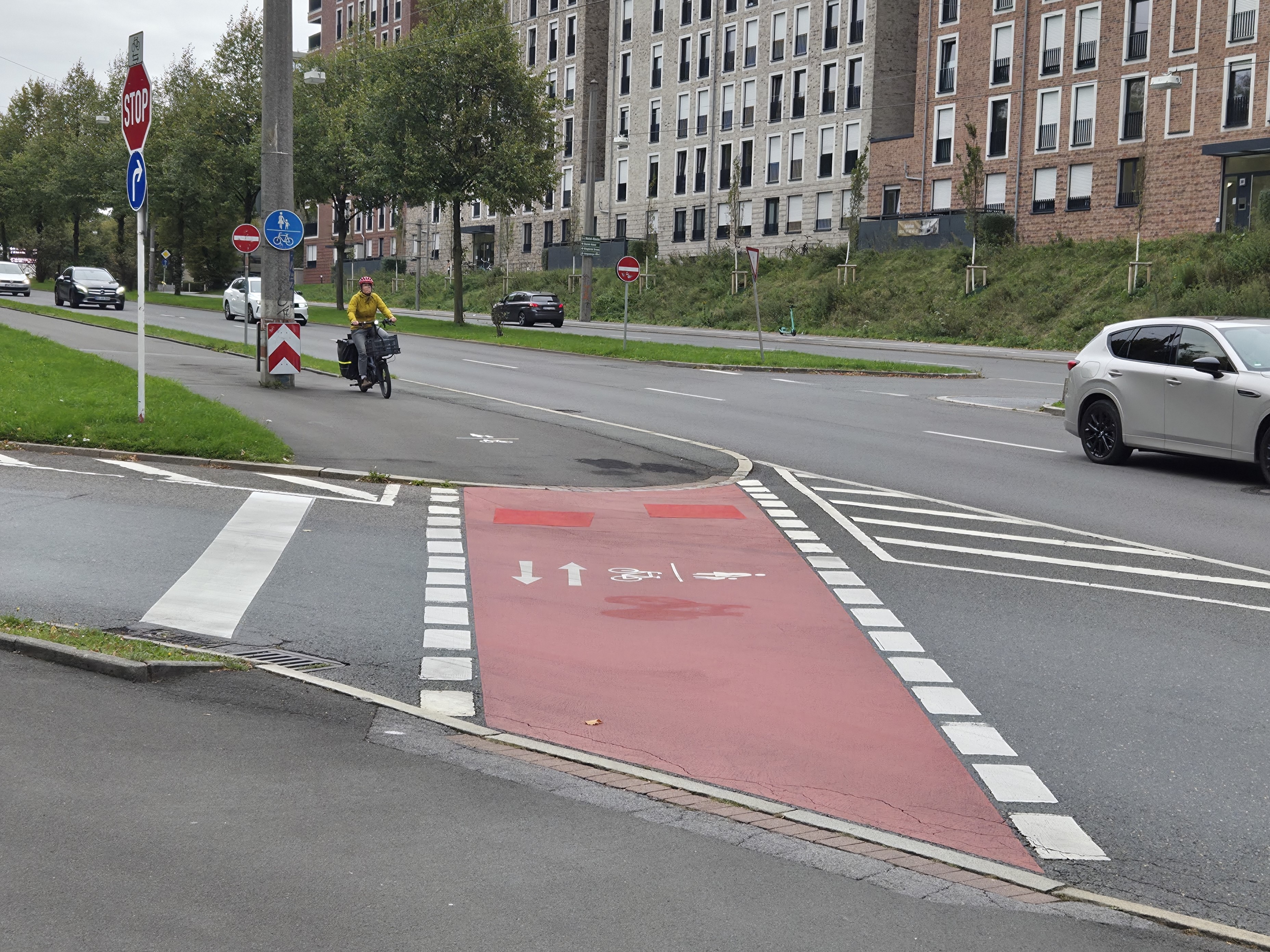 Ein roteingefärbter Geh- und Radweg, der eine einmündende Straße kreuzt. Im Hintergrund näher sich eine Radfahrerin. 