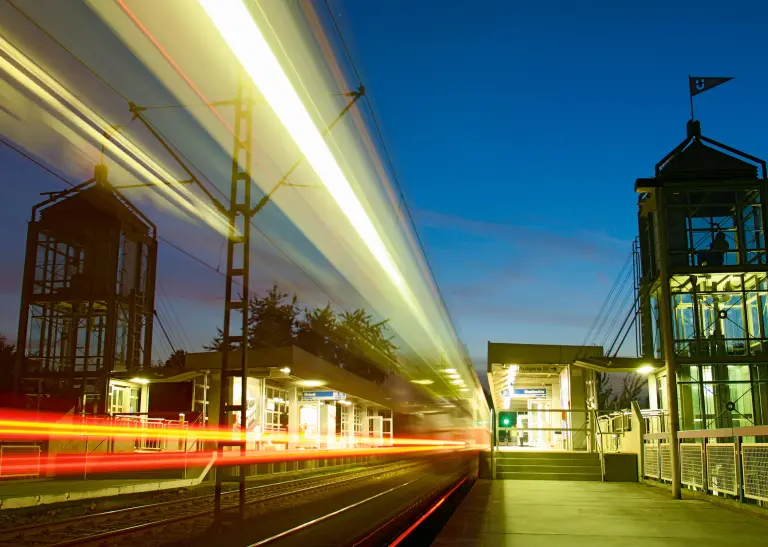 Bahnhaltestelle in der Abenddämmerung, klarer Blauer Himmel, links im Bild ist die Beleuchtung verwischt und sieht sich entlang der Gleise